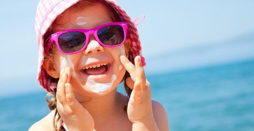 Sun Protection For Kids Young Girl Rubbing Sunscreen On Her Face, Wearing Sun Glasses And A Sun Hat