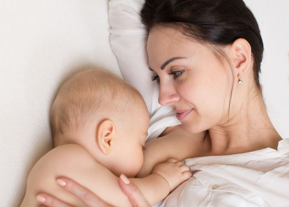 Smiling brunette mother laying on her side while breastfeeding on the bed