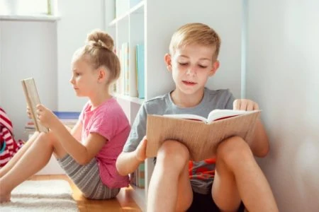 Brother and sister reading on the floor beside a bookshelf