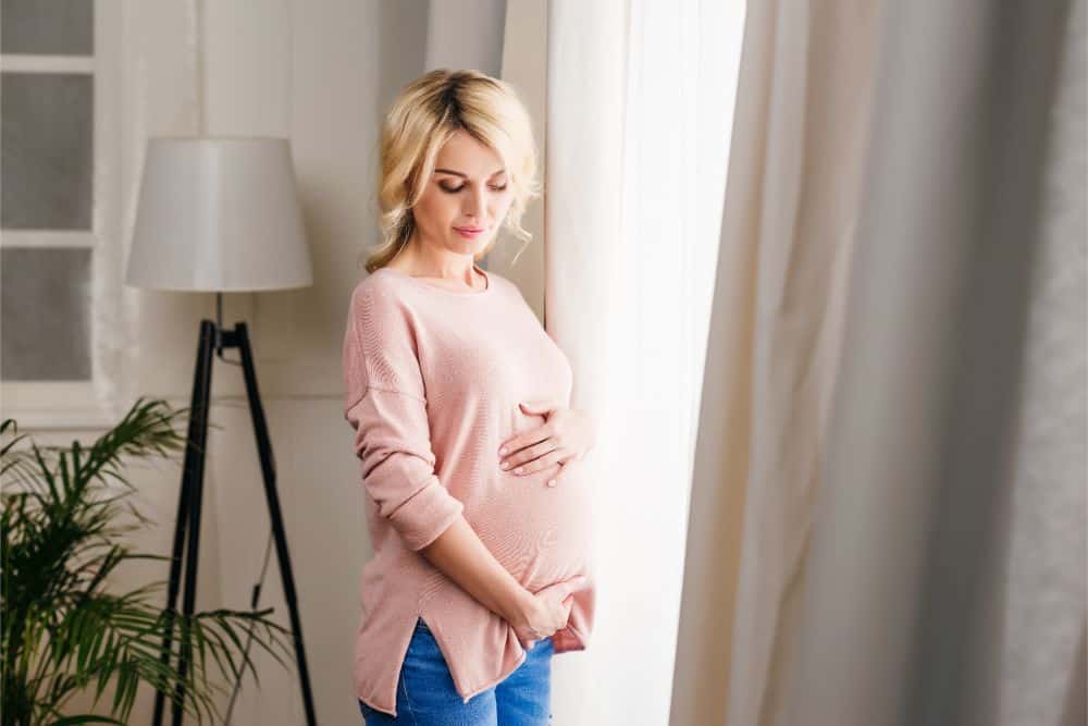 Blonde pregnant woman holding her belly in front of the window