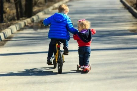 Little boy riding a bike and teaching toddler sister to ride a scooter
