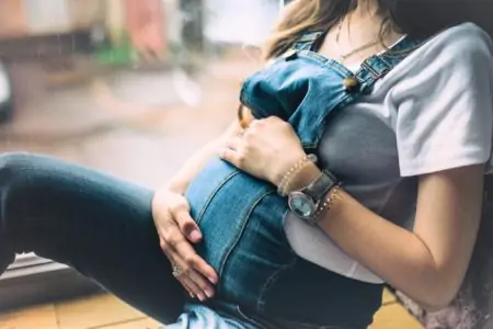 A pregnant woman, dressed in blue denim overalls, holding her hands on her tummy