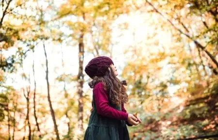 Low angle view of little toddler girl holding leaves and standing in autumn forest
