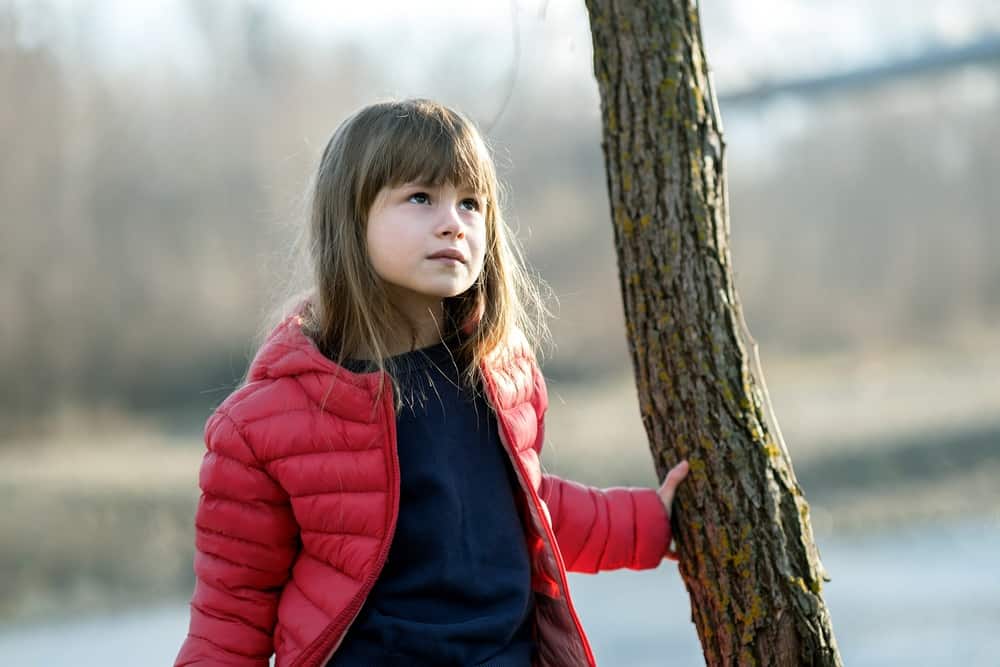 Beautiful little girl in red padded jacket standing near a tree trunk
