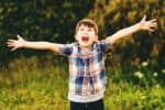 Happy kid boy having fun outdoors wearing blue plaid shirt, arms wide open