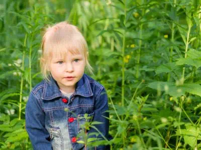 Blonde little girl dressed in blue denim suit looking out from the thick green grass