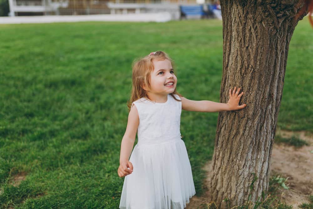 Smiling little cute girl in white dress leaning on tree in the park