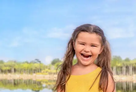 Adorable girl in yellow dress smiling brightly near the lake on sunny day