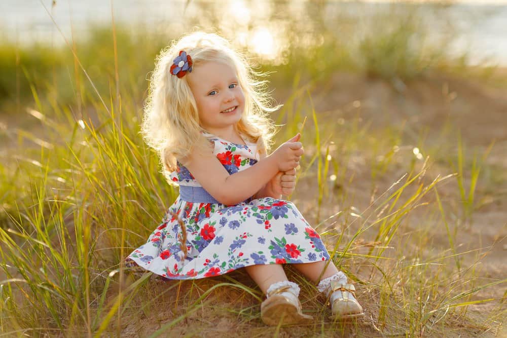 Cute little curly blonde girl sitting on sand and grass in sunny day