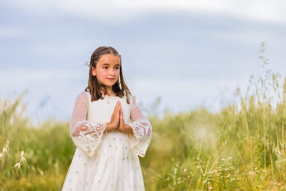 Adorable girl in white dress doing praying gesture in green meadow on sunny day