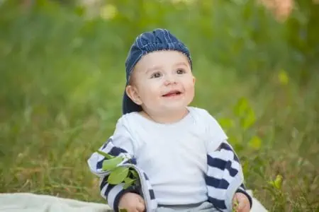 Adorable little boy wearing cap sitting on blanket on the grass looking up smiling brightly