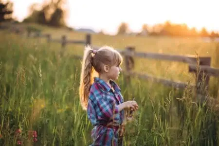 Little girl looking far away standing near wood fence in the field during sunset hours
