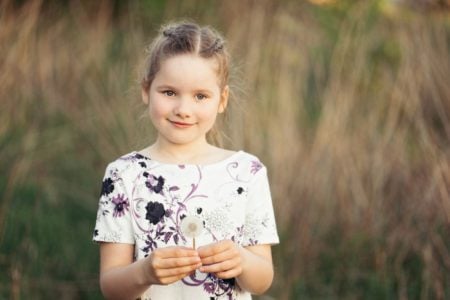 Cute young girl in white dress holding dandelion and smiling in meadow