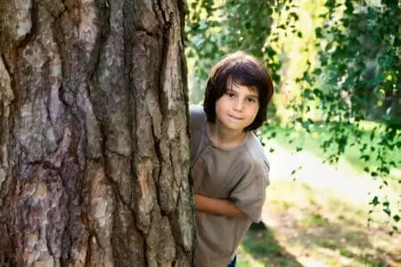 Handsome young boy peeking out from behind tree trunk in the park
