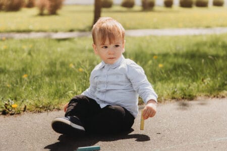 Little boy toddler holding chalk while sitting on asphalt with drawings in the park on sunny day