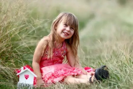Cheerful little girl in dress with small doll house sitting on the grass