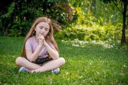 Cute young girl sitting on the grass with her eyes closed and praying
