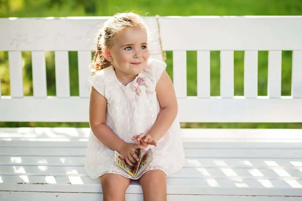 Adorable little blonde girl sitting on white bench in the park on a sunny day