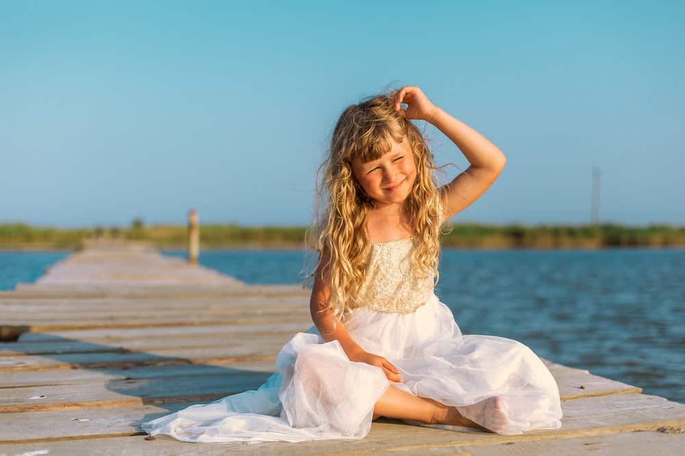 Little girl with long blond curly hair wearing white dress sitting on the pier near the se