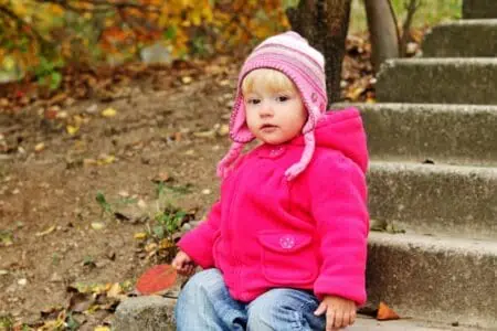 Adorable little girl wearing pink jacket and hat sitting on stairs in autumn park