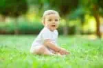 Cute toddler boy in white bodysuit sits on green summer grass