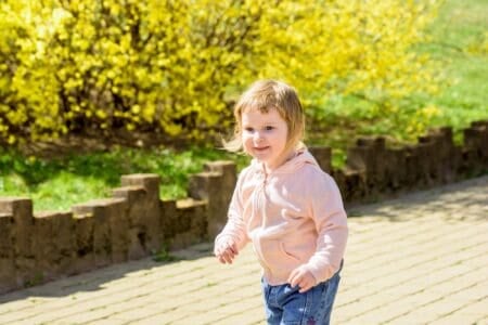 Cheerful little girl with blond hair running near blooming yellow forsythia bush