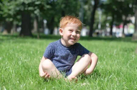 Adorable little red-haired boy sits in the park on the lawn and smiles