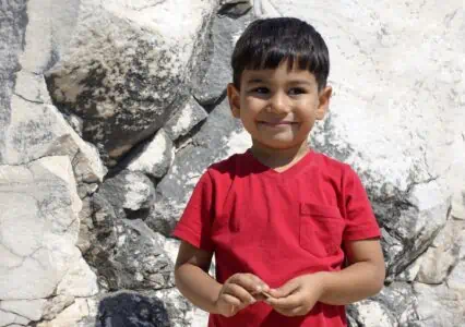 Cheerful black-haired little boy in red shirt standing against rock formation outdoors on sunny day