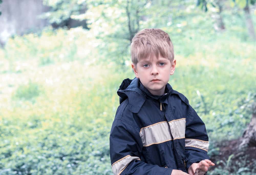 Handsome young blond boy in raincoat spending time in green forest