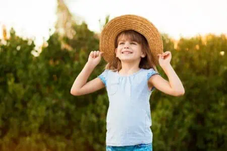 Adorable girl wearing straw hat looking up while standing in the garden on sunset day