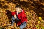 Puerto rican young boy wearing red jacket sitting on the ground full of autumn leaves