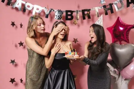 Friends holding cake and covering the eyes of birthday girl over pink wall with elegant decor