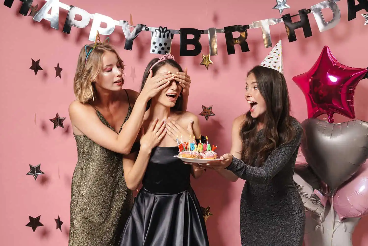 Friends holding cake and covering the eyes of birthday girl over pink wall with elegant decor