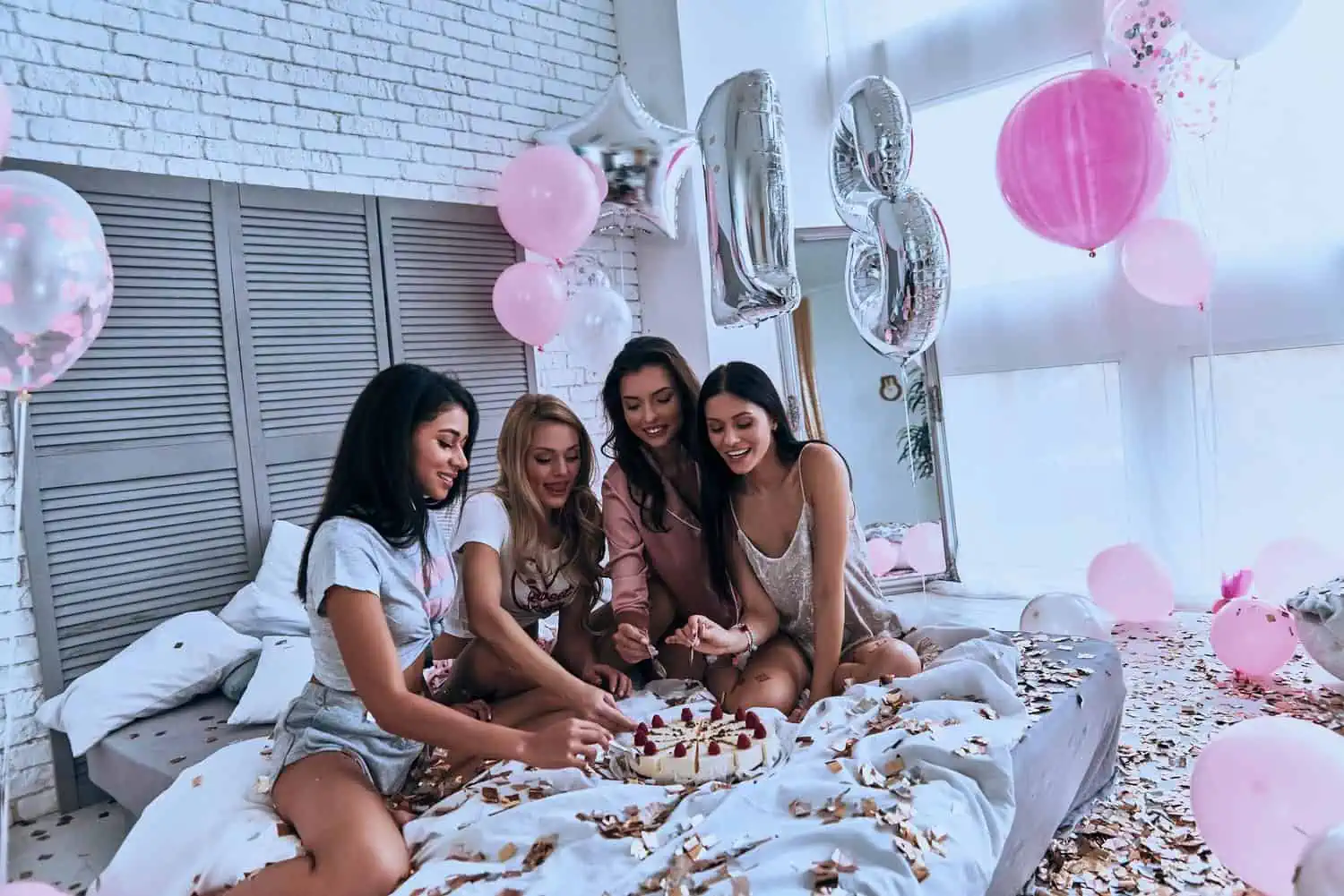 Four beautiful ladies in pajamas sitting on the bed eating cake with birthday decors all over bedroom