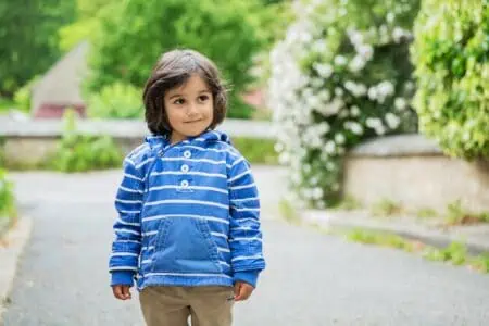 Handsome baby boy in striped blue jacket standing outdoor near flowery garden