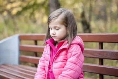 Beautiful little girl wearing pink coat sitting on wood bench at the park in autumn