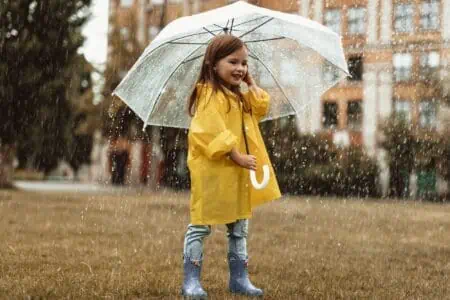 Happy girl in yellow raincoat holding clear umbrella under the rain outdoors