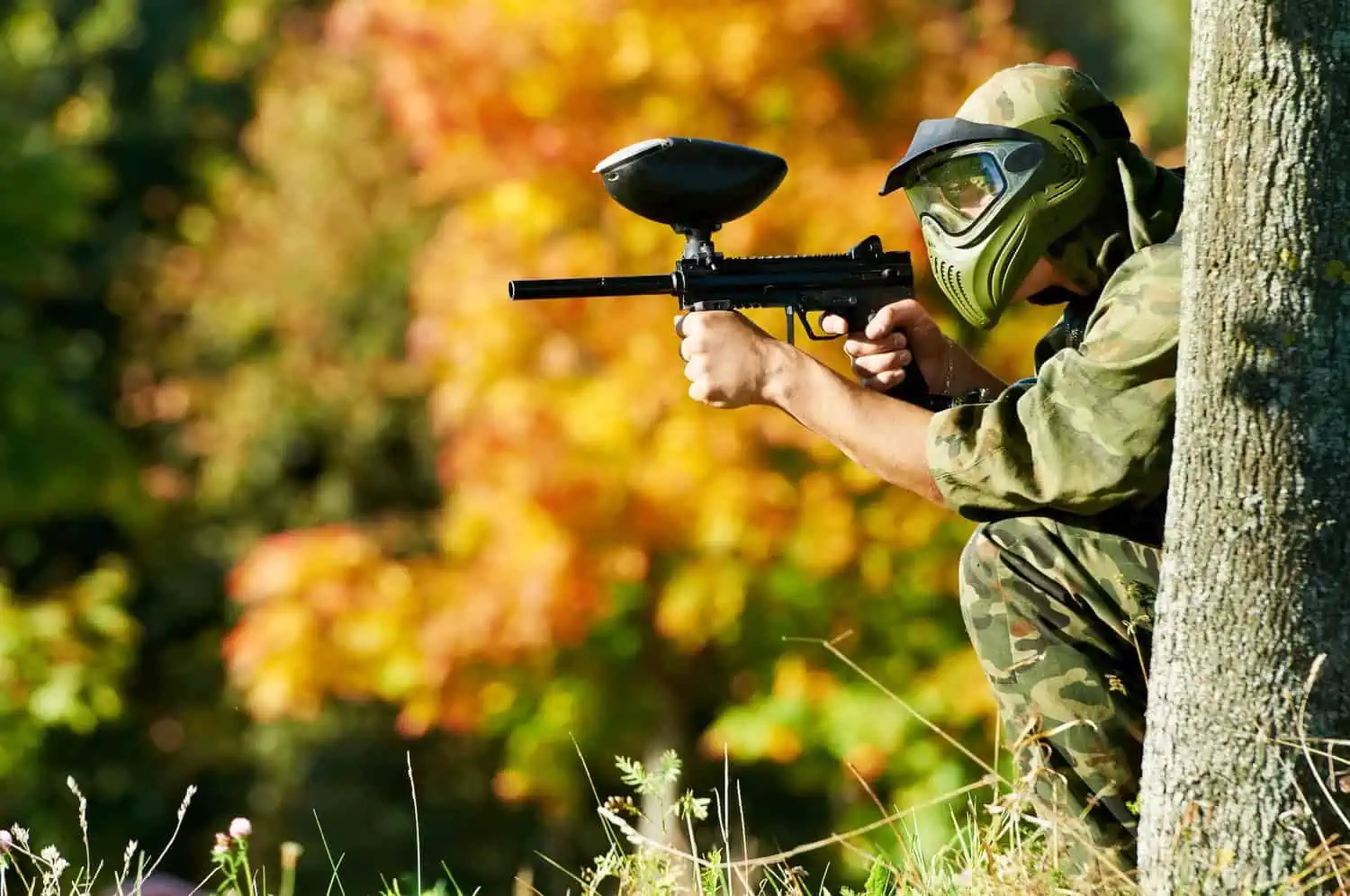 Paintball player in protective uniform and mask aiming and shooting with gun outdoors