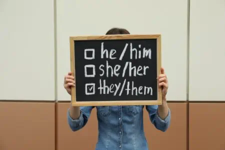 Woman in denim holding chalkboard with list of gender pronouns near color wall