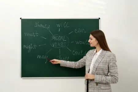 Female teacher giving lesson on modal verbs near chalkboard in classroom