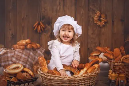Happy baby girl in chef costume sitting in front of basket full of bread