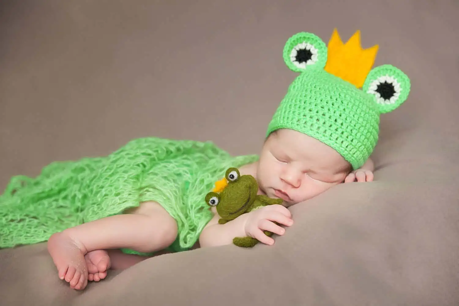 Cute newborn baby in frog costume with plush toy frog sleeping tightly on the bed