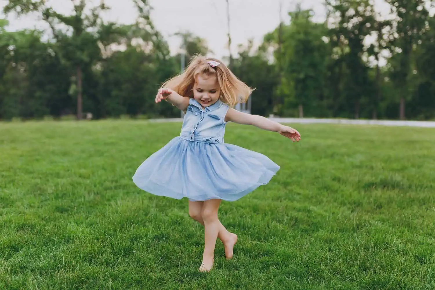Adorable baby girl in denim dress circling around on green grass law