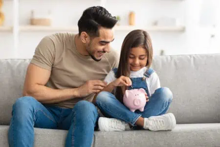 Smiling little girl putting coin in pink piggy bank, sitting with dad on the couch at home