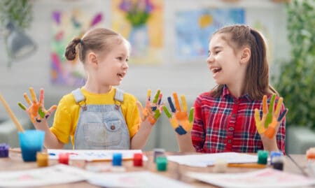 Happy young girls looking at each other with paint in their palms in front of the table with painting materials