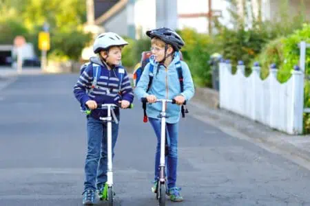 Two boys in colorful clothes riding scooter wearing safety helmets on the way to school