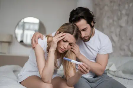 Sad husband comforting his depressed infertile wife holding negative pregnancy test on bed at home