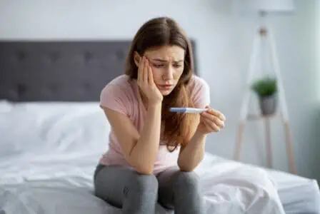 Scared young lady holding pregnancy test while sitting on bed at home