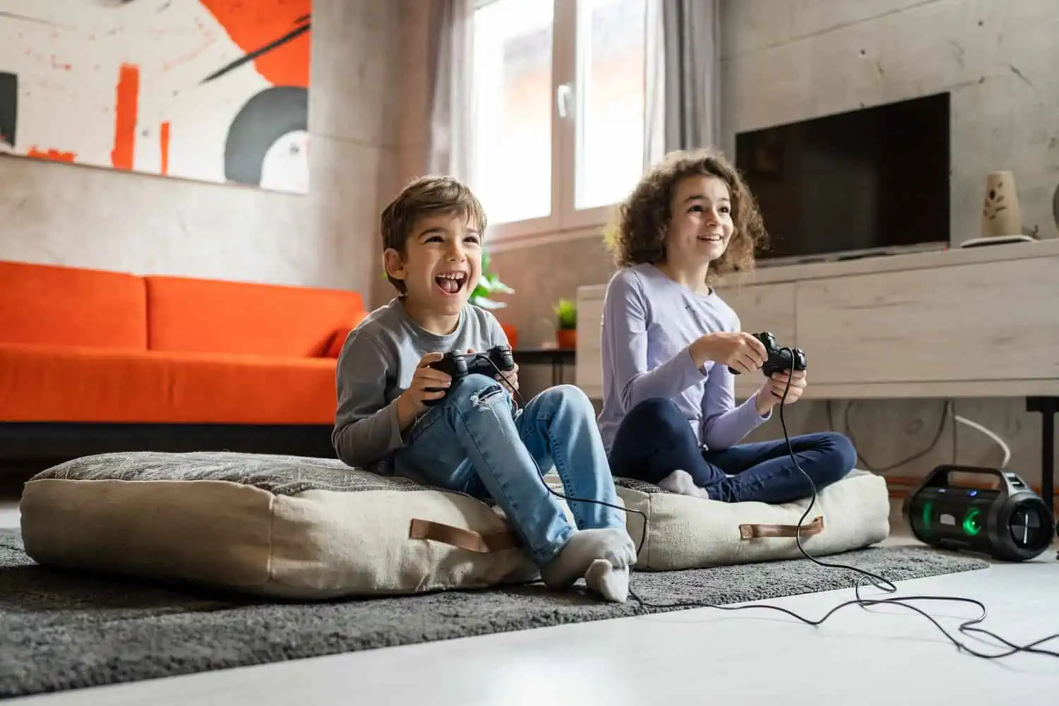 Two happy kids playing video games while sitting on living room floor at home