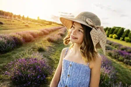 Blonde girl wearing summer hat standing in lavender field during sunset